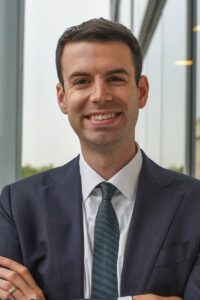 A smiling man with short dark hair wearing a dark suit, white shirt, and dark tie, standing with his arms crossed in front of a large window with a view of greenery outside.