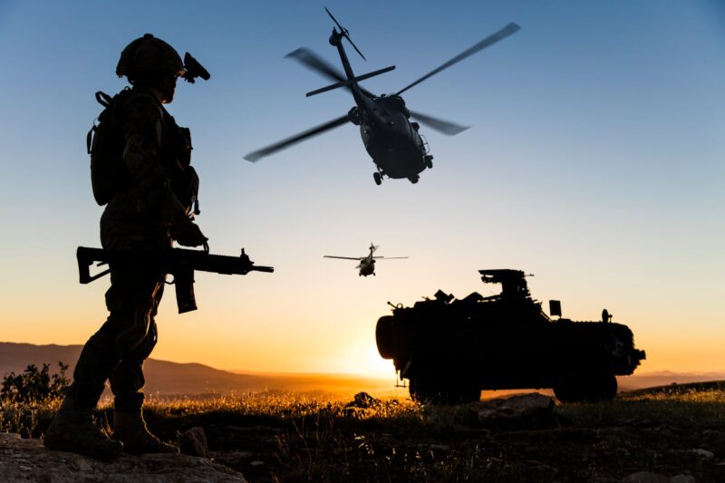 A soldier in combat gear silhouetted at sunset beside an armored military vehicle as helicopters fly overhead